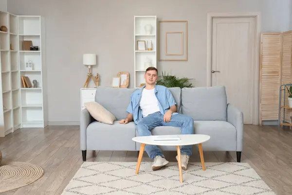 Genç adam oturma odasındaki gri kanepede dinleniyor.