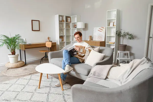 Genç adam oturma odasındaki gri koltukta dergi okuyor.