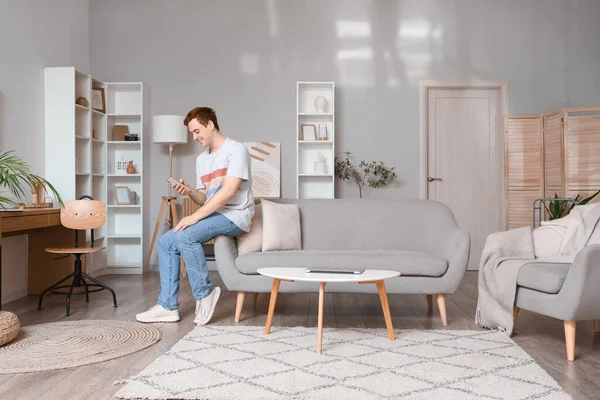 Genç adam oturma odasındaki gri koltukta cep telefonu kullanıyor.