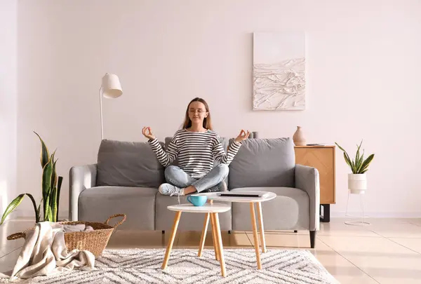 Gözlüklü genç kadın evdeki gri kanepede meditasyon yapıyor.