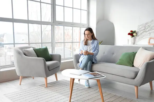 Oturma odasındaki gri kanepede çay içen güzel genç bir kadın.