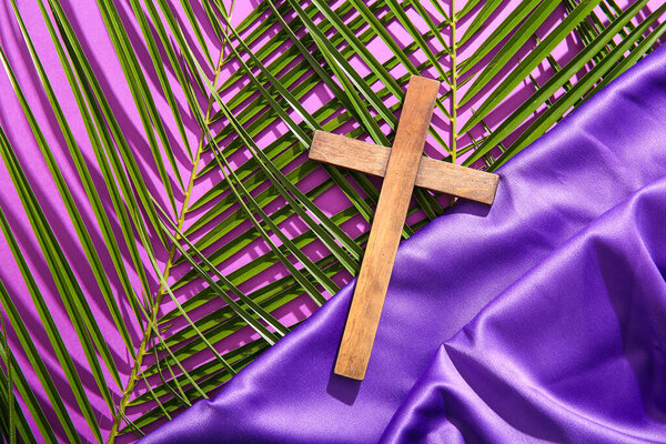Wooden cross with palm leaves and purple fabric on color background