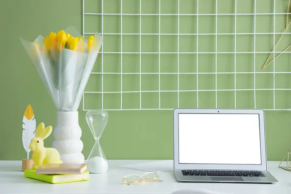 Blank laptop, vase with tulips and Easter rabbit on table near green wall
