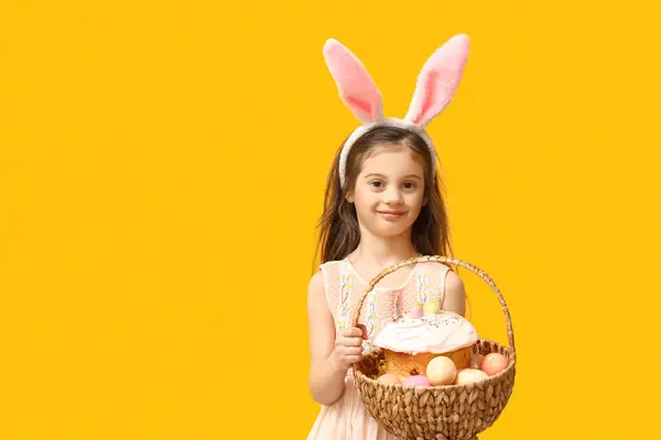 黄色の背景にイースターケーキと卵のバスケットとバニーの耳にかわいい少女 — ストック写真