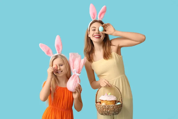 可爱的小女孩 戴着礼物蛋 她的妈妈长着兔子耳朵 手里拿着蓝底复活节蛋糕篮子 — 图库照片