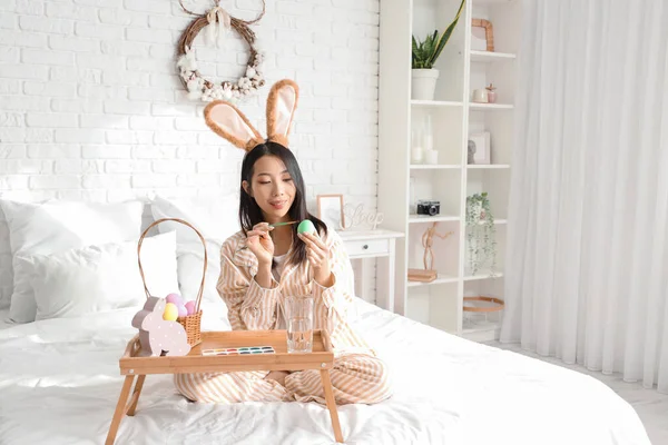 Tavşan kulaklı genç Asyalı kadın yatak odasında paskalya yumurtası boyuyor.