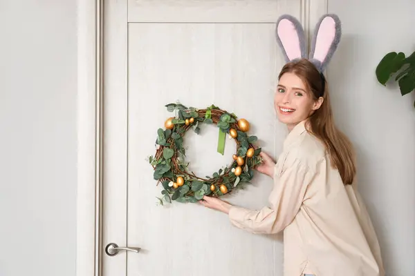 Young woman in bunny ears hanging Easter wreath on door at home