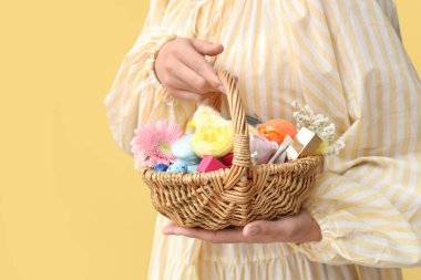 Sarı arka planda kozmetik ürünleri olan hasır bir Paskalya sepeti tutan genç bir kadın.