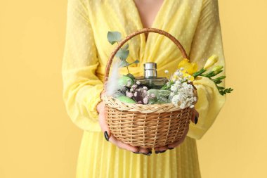 Sarı arka planda kozmetik ürünleri olan hasır bir Paskalya sepeti tutan genç bir kadın.