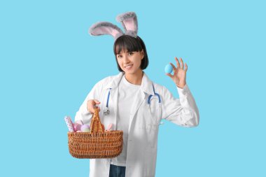 Mavi arka planda Paskalya yumurtaları olan tavşan kulaklı kadın doktor.