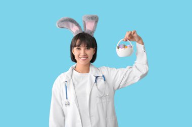 Mavi arka planda Paskalya yumurtaları olan tavşan kulaklı kadın doktor.