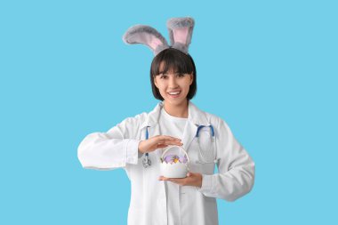 Mavi arka planda Paskalya yumurtaları olan tavşan kulaklı kadın doktor.