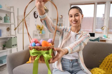 Paskalya yumurtası sepeti olan genç bir kadın evdeki kanepede oturuyor.