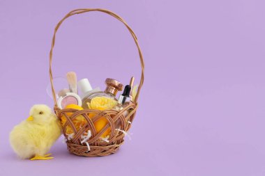 Dekoratif kozmetik ürünleri, paskalya yumurtaları ve mor arka planda bebek tavuğu olan hasır sepet.
