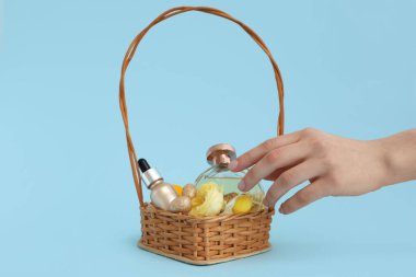 Dekoratif kozmetik ürünlerle dolu hasır sepetli kadın elleri, mavi arka planda Paskalya yumurtaları ve çiçekler