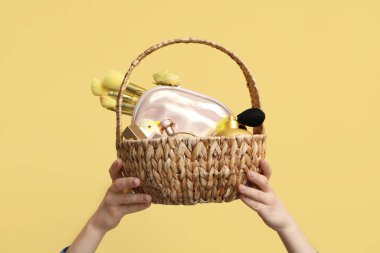 Hasır sepeti dekoratif kozmetiklerle dolu kadın elleri ve sarı arka planda bebek tavuğu.