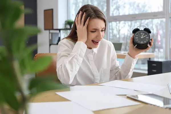Stressed young businesswoman with alarm clock at table in office. Time management concept