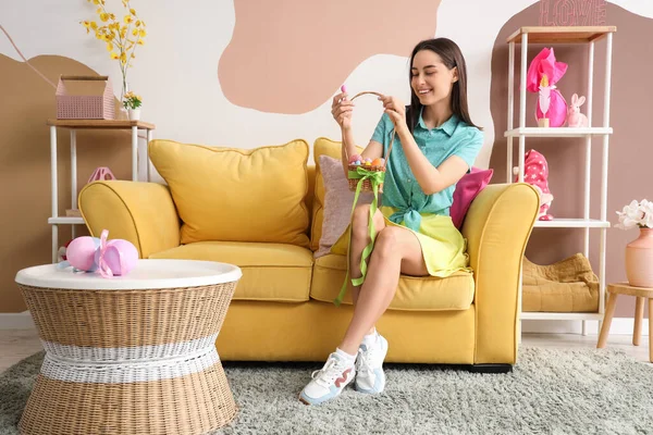 Paskalya yumurtası sepeti olan genç bir kadın evdeki kanepede oturuyor.