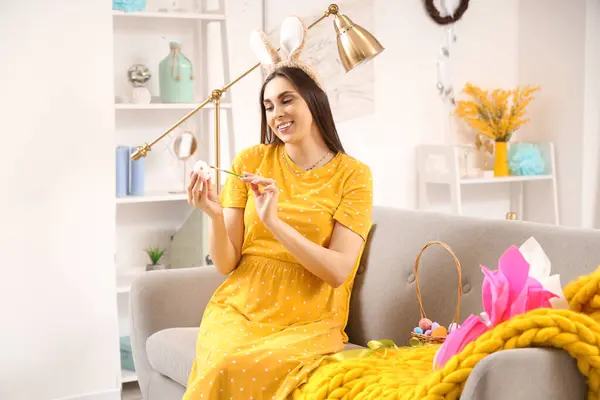 Young pregnant woman in bunny ears painting Easter egg at home
