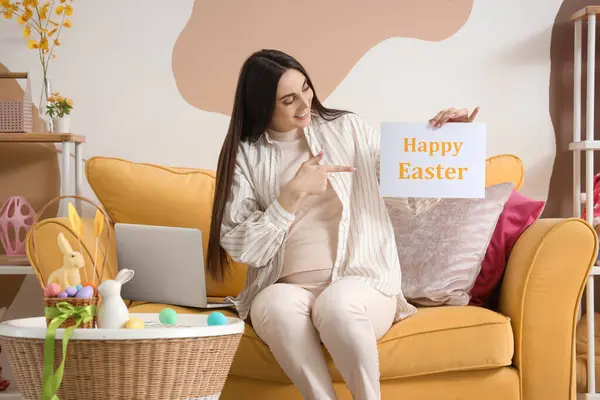 Young pregnant woman holding paper with text HAPPY EASTER at home