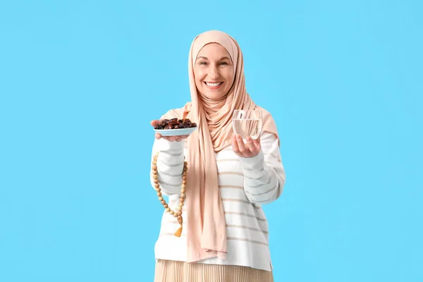 Mature Muslim woman with dates, glass of water and praying beads on blue background. Ramadan celebration