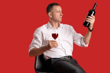 Kırmızı arka planda oturan cam ve şişe şarabı olan olgun bir adam.