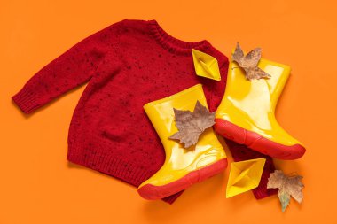 Süveterli sarı çizmeler, kağıt gemiler ve turuncu arka planda sonbahar yaprakları.