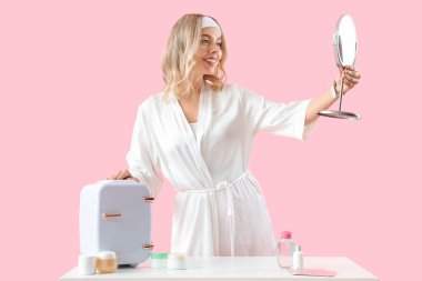 Kozmetik ürünleri için küçük buzdolabı olan güzel, genç bir kadın pembe arka planda.