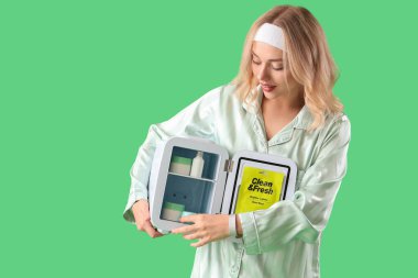 Yeşil arka planda küçük kozmetik buzdolabı olan güzel, genç, mutlu bir kadın.