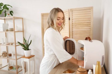 Güzel, genç, mutlu bir kadın banyodaki küçük buzdolabından kozmetik ürünler alıyor.