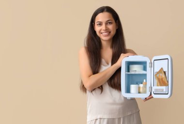 Bej arka planda küçük kozmetik buzdolabı olan güzel, genç, mutlu bir kadın.