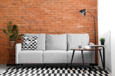 Kahverengi tuğla duvarın yanında rahat gri kanepesi ve sehpası olan modern oturma odasının içinde.