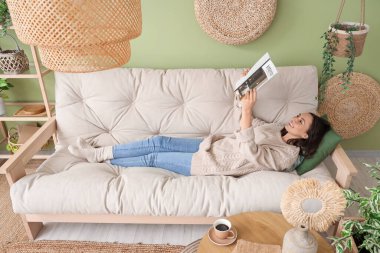 Elinde dergi olan güzel bir kadın oturma odasındaki rahat beyaz koltukta dinleniyor.