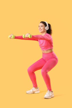 Kulaklıklı, sarı arka planda dambılları olan genç bir kadın.