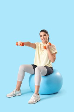 Mavi arka planda, halterli, spor yapan genç bir bayan.