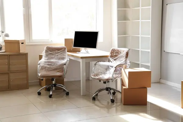 Interior of office with chairs wrapped in stretch film and cardboard boxes on moving day