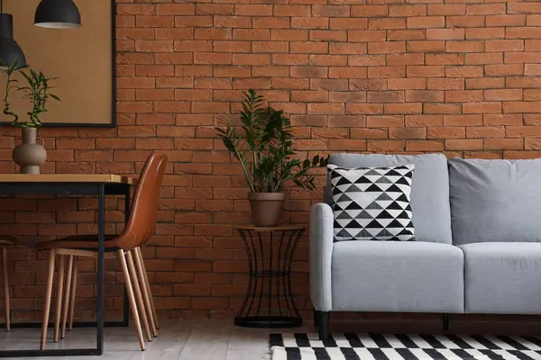 Interior of modern living room with cozy grey sofa and dining table near brown brick wall