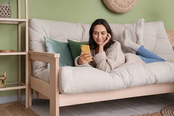 Oturma odasındaki rahat beyaz koltukta dinlenen cep telefonlu güzel genç bir kadın.