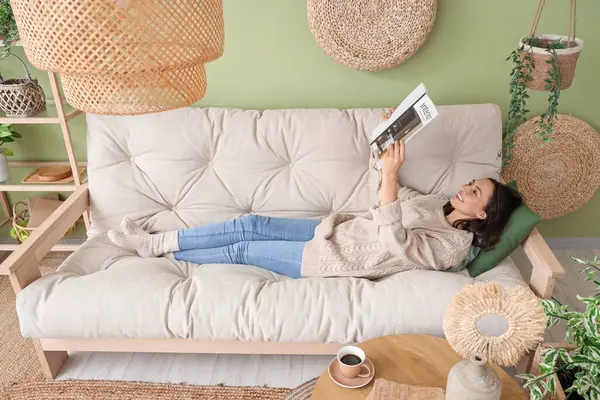 Elinde dergi olan güzel bir kadın oturma odasındaki rahat beyaz koltukta dinleniyor.
