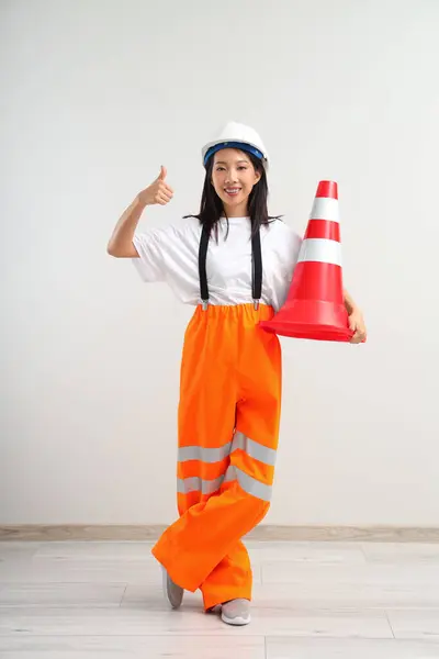 Asian woman in jumpsuit and hardhat with traffic cone near white wall