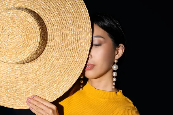 Koyu arkaplanda güzel küpeleri ve şapkası olan genç Asyalı kadın.