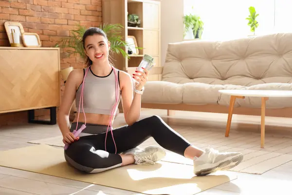 Sporty young woman with skipping rope and water bottle at home