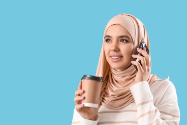Elinde kahve fincanı olan genç Müslüman kadın mavi arka planda cep telefonuyla konuşuyor, yakın plan.