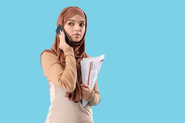 Gazeteyle tesettürlü genç Müslüman kadın mavi arka planda cep telefonuyla konuşuyor