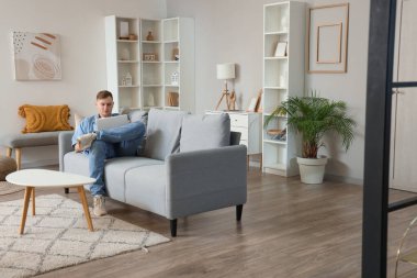 Genç adam oturma odasındaki gri koltukta dizüstü bilgisayar kullanıyor.