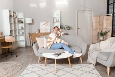 Genç adam oturma odasında gri koltukta oturuyor.