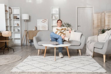 Genç adam oturma odasında gri koltukta oturuyor.