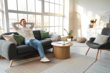 Genç adam oturma odasındaki pencerenin yanındaki yumuşak kanepede dinleniyor.