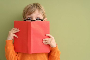 Gözlüklü, renkli arka plan kitaplarıyla yüzünü kapatan küçük bir çocuk.