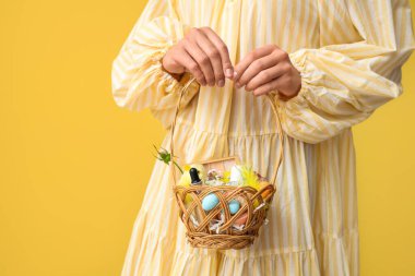 Sarı arka planda kozmetik ürünleri olan hasır bir Paskalya sepeti tutan genç bir kadın.